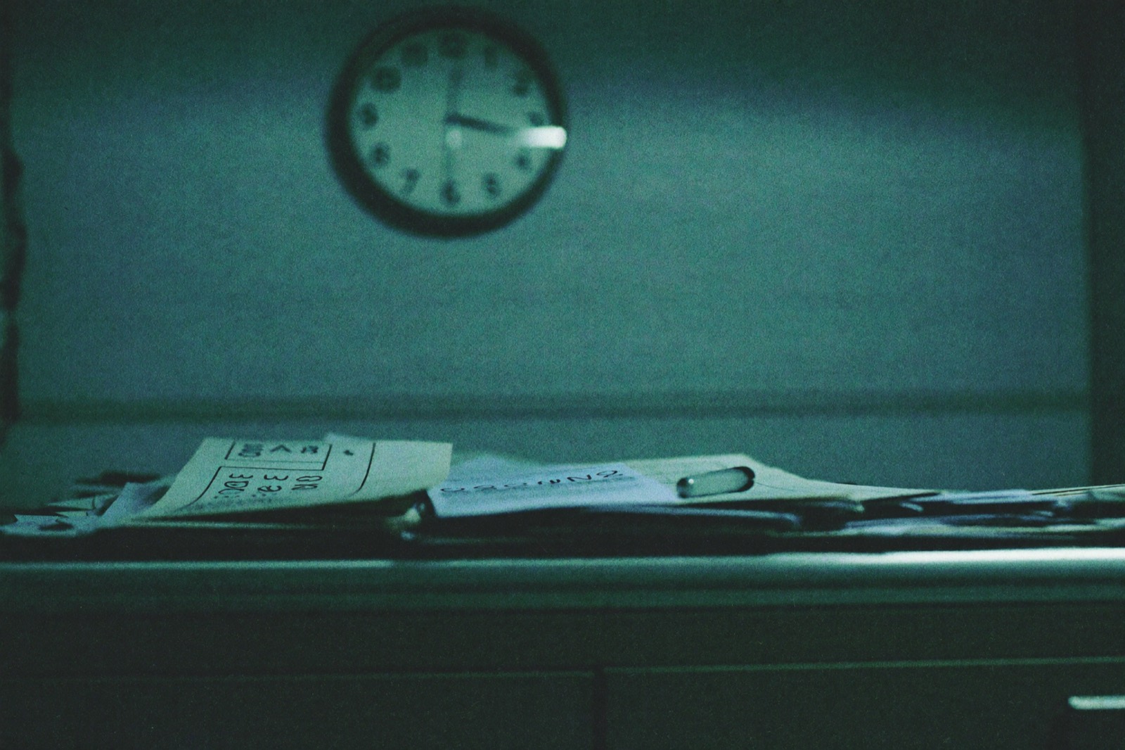 Mesa de hospital com fichas de papel acumuladas sob a luz fria, relógio de parede desfocado ao fundo