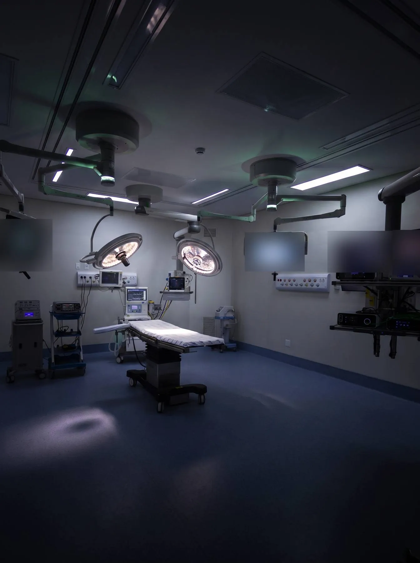 Sala cirúrgica moderna com focos cirúrgicos acesos, mesa ao centro e monitores nas paredes, em luz teal